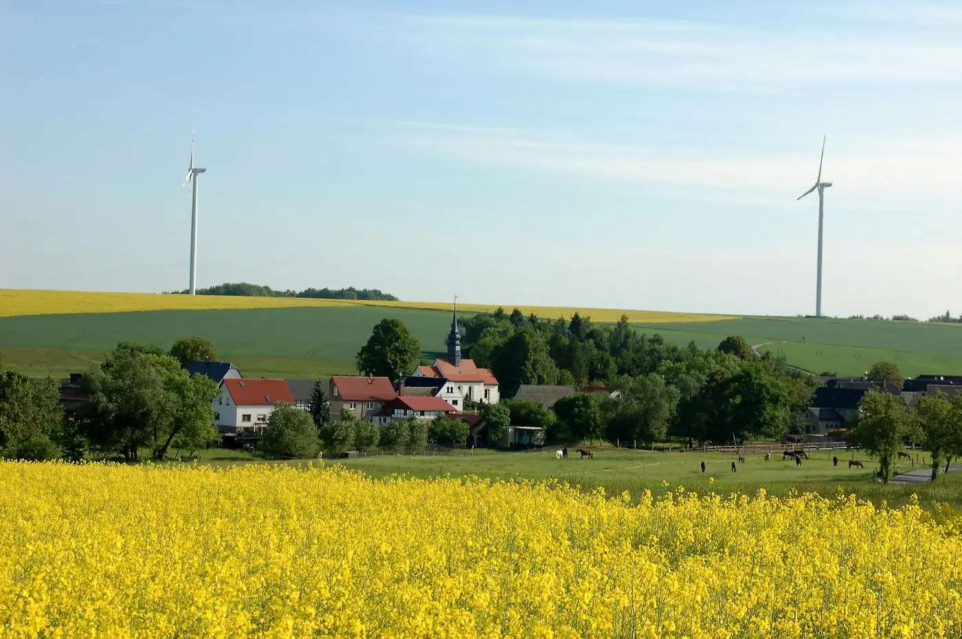 Dorf mit Kirchturm hinter einem blühenden Rapsfeld, Windräder am Horizont Dorf mit Kirchturm hinter einem blühenden Rapsfeld, Windräder am Horizont