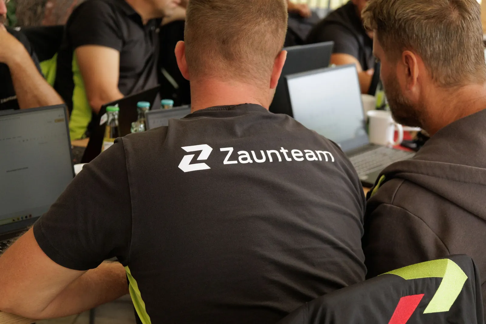 Mitarbeiter im Zaunteam-Shirt von hinten, mit Kollegen an Laptops am Besprechungstisch Mitarbeiter im Zaunteam-Shirt von hinten, mit Kollegen an Laptops am Besprechungstisch